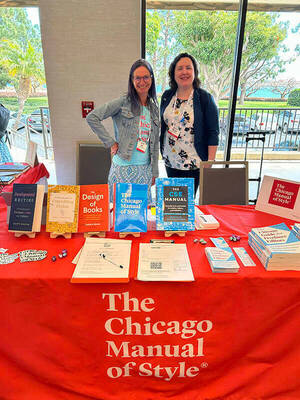 Two women pose for a photo behind a table draped in red cloth with the Chicago Manual of Style logo on it.
