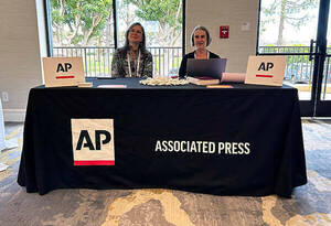Two women sit behind a table draped in a block cloth with the Associated Press logo on it.