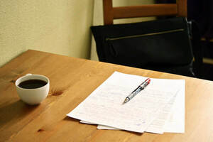 A red pen rests on a stack of papers next to a full mug of black coffee on a wooden desk.