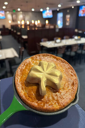 A photo of a green skillet with tan cornbread and a gold pat of butter on top. The skillet is in the foreground and the background is a blurred image of the restaurant.