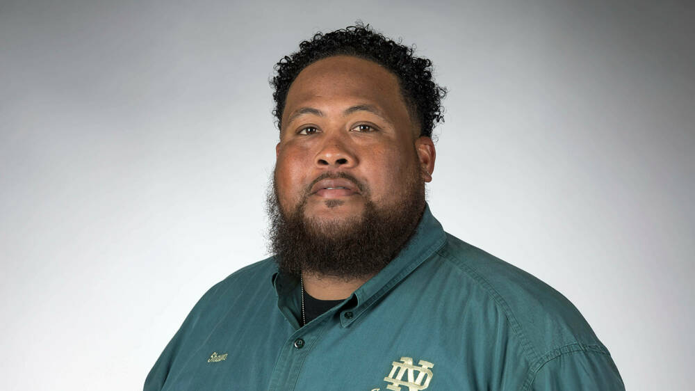 A man with brown skin and short black hair, with a black beard, poses for a portrait. He is wearing a green button down shirt.