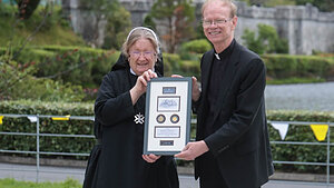 Smiling nun and Rev. Robert K. Dowd, C.S.C., holding framed plaque outside Kylemore Abbey with green hills.